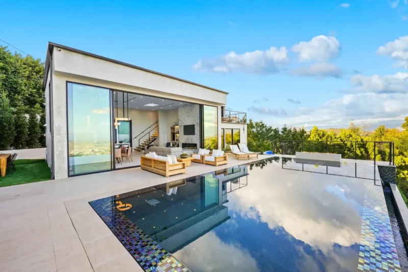 Glass-walled pool house beside a dark tiled infinity pool on a hillside deck