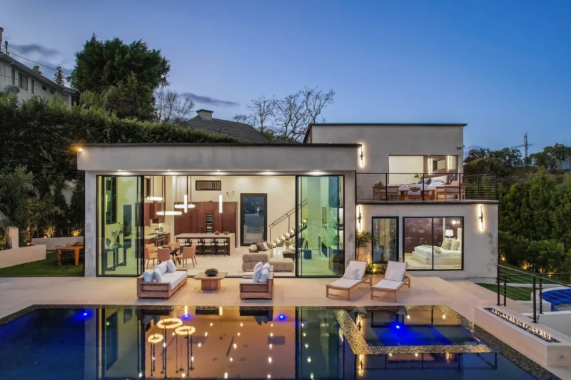 Twilight aerial of the lit glass-walled home above a blue-lit pool
