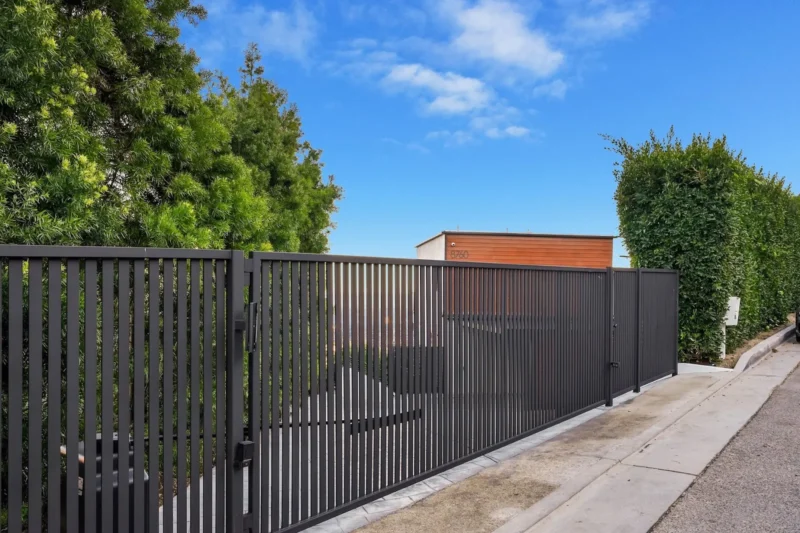 Dark slatted metal gate and fence along a concrete walkway beside a hedge